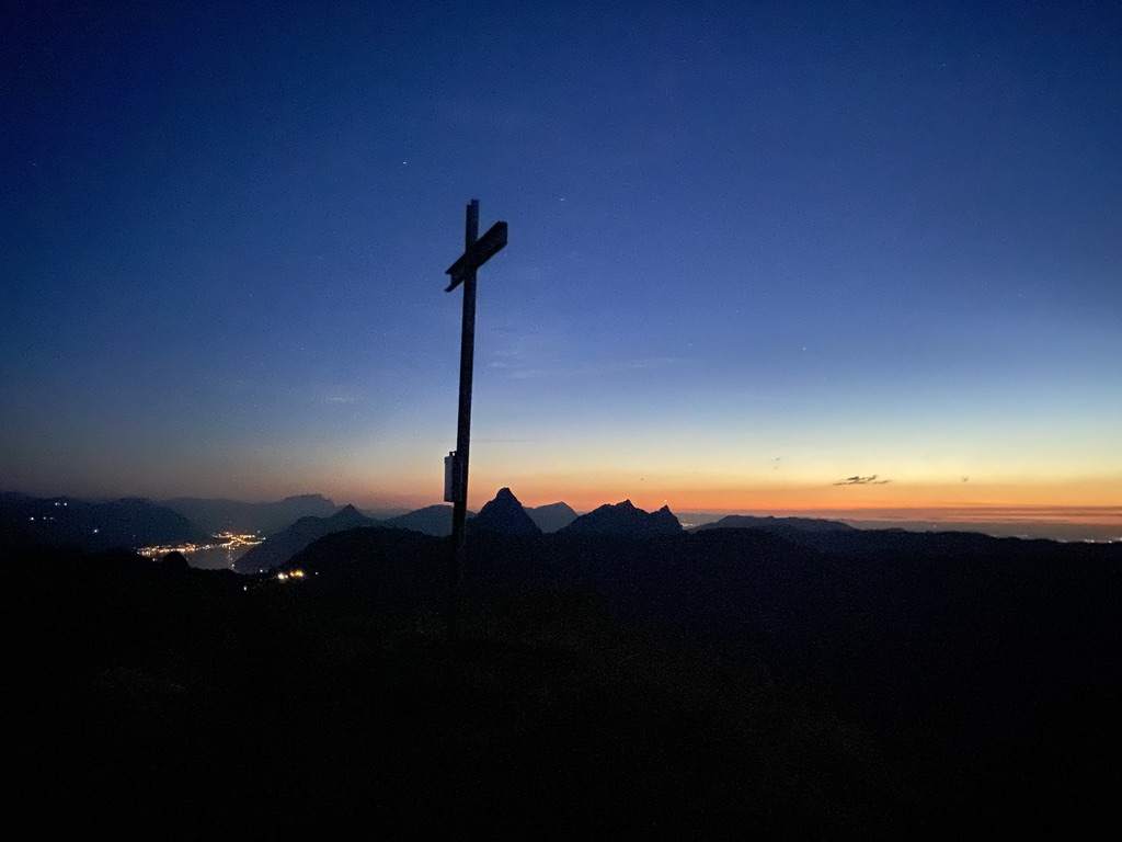 Gipfelkreuz mit Sonnenaufgang und mehreren Bergipfeln blauem Himmel und Umrissen